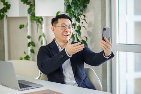 Millennial asian man using smartphone for online video conference call waving hand making hello gesture on office.の写真素材