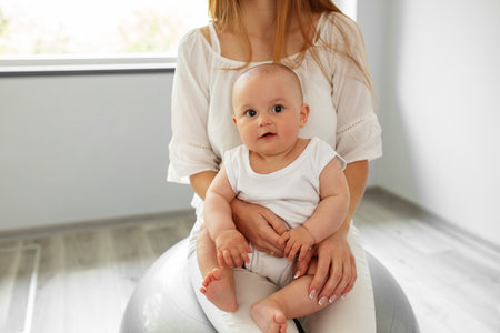 Happy mother holding baby girl while bouncing on fitness ball in cozy home. Stability ball exercises for neurodevelopment of baby.の写真素材