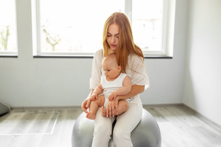 Happy mother holding baby girl while bouncing on fitness ball in cozy home. Stability ball exercises for neurodevelopment of baby.の写真素材