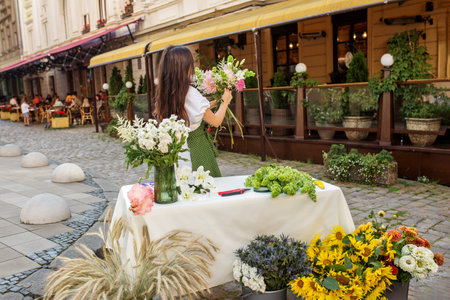 Focused florist in green polka dot apron arranges colorful selection of flowers at her charming street-side standの写真素材