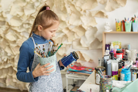 Concentrated young girl selects paintbrush, ready to create, amidst vibrant array of paints and art toolsの写真素材