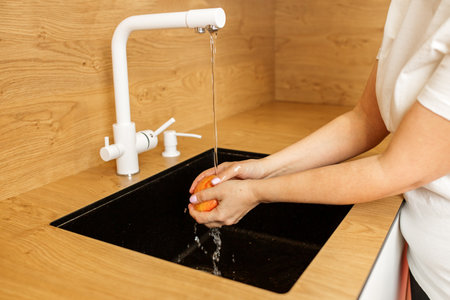 Hands are seen rinsing red apple under flowing water from white tap over black kitchen sink, ensuring fruit is clean for eatingの写真素材
