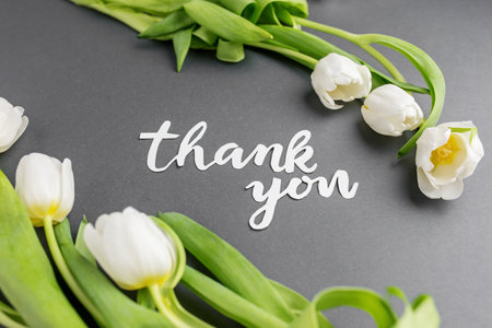Beautiful arrangement of white tulips next to 'thank you' message in elegant white script on contrasting grey background.の写真素材
