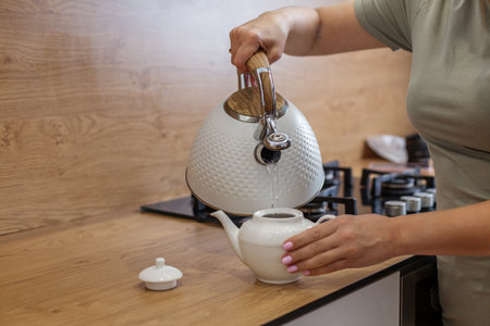 Close-up of a woman's hand pouring water from textured white kettle into ceramic teapot on wooden...