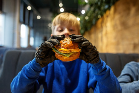 Preteen child boy in blue sweater, wearing black gloves, holds up a gourmet burger, ready to eat in cozy restaurant setting.の写真素材