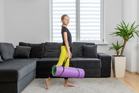 Young girl with ponytail walks with purple yoga mat in home environment, preparing for fitness routine.の写真素材
