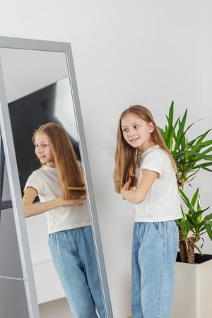 Smiling preteen child brushes her long hair while looking at her reflection in full-length mirror in bright room.の写真素材