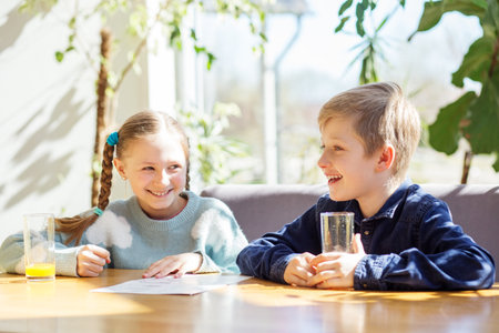 Brother and sister smiling and looking at each other while reading a menu at a wooden table, basking in natural light, with plants in background.の写真素材