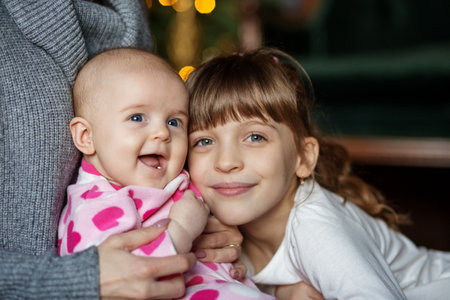 An older sister gently hugs her baby sister, both smiling joyfully in cozy, warm indoor environment.の写真素材