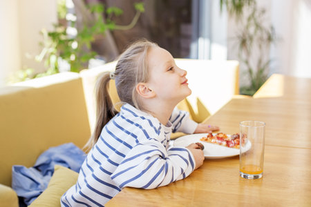Kid leaning forward and smiling while eating slice of pizza at sunny indoor table with glass of orange juice.の写真素材