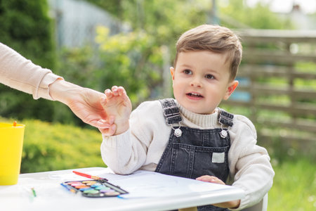 Toddler boy is enjoying painting activity at small table in garden, guided by parent's hand on sunny...