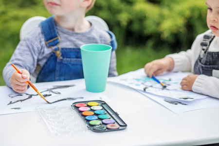 Close-up of two young children engaged in painting with watercolors at table outdoors, enjoying creative playtime.の写真素材