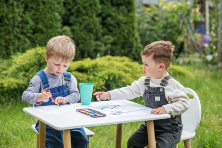 Two young boys sit at table in backyard, painting with watercolors and enjoying creative outdoor...