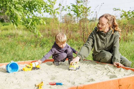 Mother and young son enjoy sunny day playing with toy trucks in sandbox at an outdoor park.