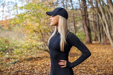 Blonde woman wearing black activewear and cap stands confidently in colorful autumn forest, hands on hips.の写真素材