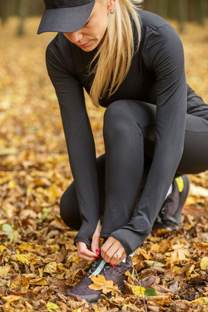Close-up of blonde woman in black sportswear tying her shoe while crouching among fallen autumn leaves in forest.の写真素材