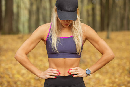 Close-up of fit blonde woman wearing sports bra and cap, standing confidently in an autumn forest after workout.の写真素材