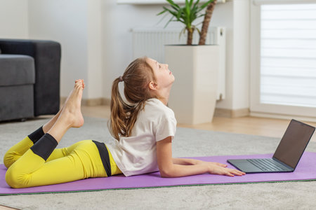 Child following an online yoga class while performing backbend on yoga mat at home, focusing on flexibility and posture during practice.の写真素材