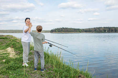 Mother gives son high-five after successful fishing moment by lake, enjoying quality time outdoors together.の写真素材