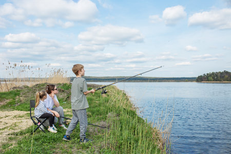 Mother and two children enjoy relaxing fishing trip by riverbank, surrounded by nature and clear skies.の写真素材