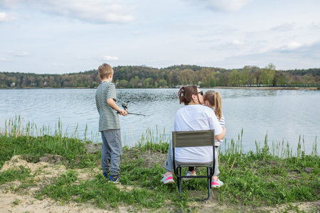 Mother kisses daughter while son fishes nearby on peaceful shore of lake, enjoying calm day outdoors.の写真素材