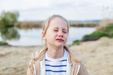 Young girl with tears in eyes stands outdoors near calm lake, visibly upset and emotional on...
