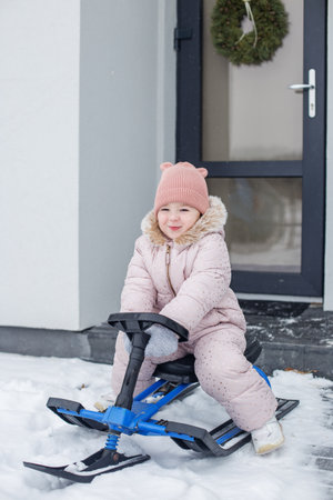 Cheerful toddler dressed in cozy jacket and hat plays outside in snow, happily riding sled near contemporary house decorated for winter.の写真素材