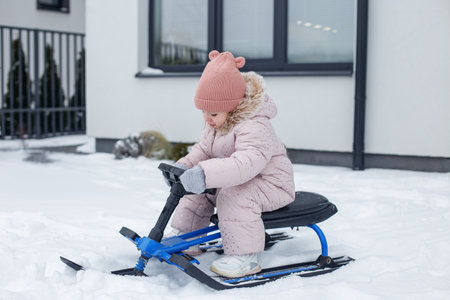 Kid dressed warmly in pink coat and hat is sitting on sled, ready to glide through soft snow in front of stylish house during winter.の写真素材