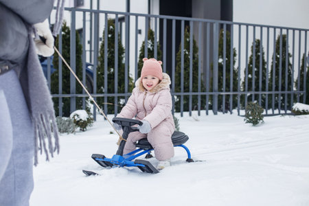 Toddler in a cozy winter outfit enjoys riding a snow sled while being towed by a parent on a snowy day in a suburban area, surrounded by white snow and greenery.の写真素材