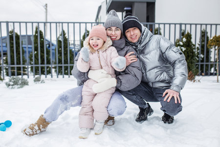 A happy family plays in the snow, bundled in warm attire. The parents kneel beside their young daughter, who joyfully poses for the camera. Their home adds a cozy backdrop.の写真素材
