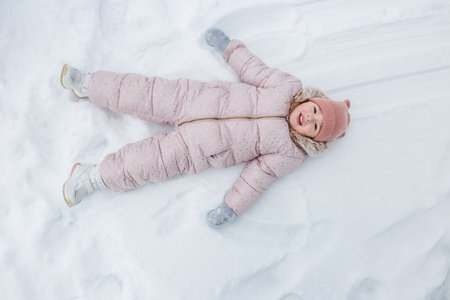 A cheerful child dressed in a pink winter coat and hat lies on the snow, moving arms and legs to create snow angels. The bright white snow surrounds them, showcasing a playful winter atmosphere.の写真素材