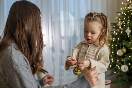 The mother and daughter joyfully decorate a Christmas tree, with lights twinkling and a warm atmosphere. The little girl is focused on hanging an ornament while her mother enjoys the moment.の写真素材