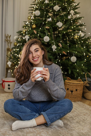 A young woman sits cross-legged on the floor, holding a warm drink. The festive Christmas tree behind her is adorned with lights and ornaments, creating a cozy atmosphere.の写真素材