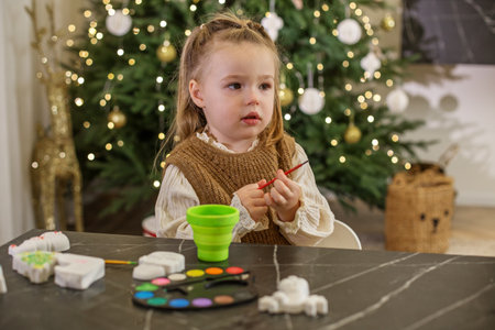 Little child sits at table, focused on their Christmas craft project. Surrounded by decorations, festive spirit fills room as colorful materials are used to create holiday decorations.の写真素材