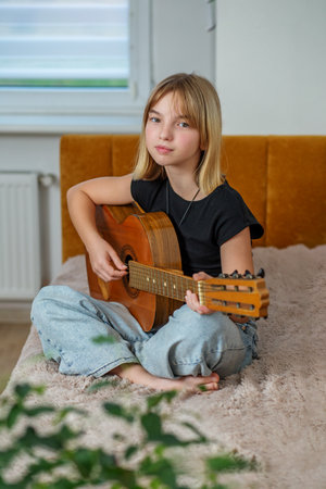 Tween sits cross-legged on cozy bed, strumming guitar while enjoying peaceful afternoon indoors.の写真素材