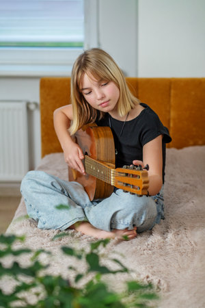 Tween sits cross-legged on bed, skillfully playing guitar in bright and inviting room. Soft light filters through window.の写真素材