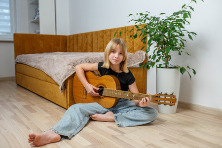 A young girl sits cross-legged on the floor, strumming her guitar in a bright, inviting space. A cozy sofa and plant add warmth.の写真素材