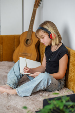 Tween girl sits comfortably on soft rug, focused on writing music in notebook. She wears headphones, and an acoustic guitar rests on wall behind her, creating creative atmosphere.の写真素材