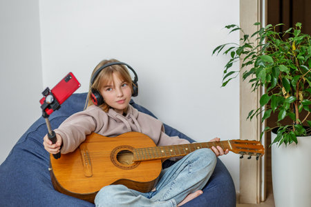 Preteen girl sits comfortably on bean bag chair, holding guitar and smartphone on selfie stick.の写真素材