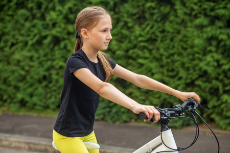 Preteen girl is confidently riding her bicycle along quiet street. She is dressed in black shirt and bright yellow pants, enjoying sunny day with backdrop of lush greenery.の写真素材