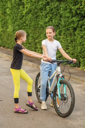 Two preteen girls are having fun in park. One girl is standing near bicycle, while other girl, wearing sporty clothes, holds her hand. Sun shines brightly, adding warmth to day.の写真素材