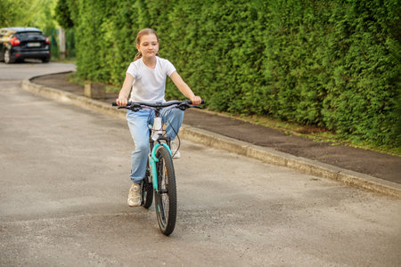 A girl, around preteen age, rides her bicycle down a peaceful suburban road lined with vibrant green hedges. She enjoys the sunny day, showcasing her carefree spirit.の写真素材