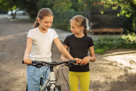 Two girls are outdoors, standing next to a bicycle in a park. One girl is adjusting the bicycle brakes while the other watches, both are smiling and enjoying the moment, surrounded by trees.の写真素材