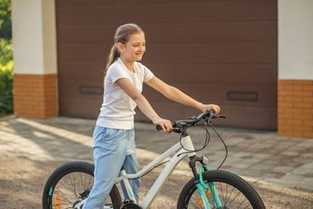 A girl smiles as she stands next to her bicycle in a peaceful residential neighborhood. The sun shines brightly, and she is dressed casually in a t-shirt and sweatpants, ready for a ride.の写真素材