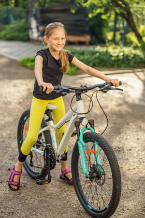 A girl with long hair is smiling while standing next to her bicycle in a park. She wears black and yellow athletic clothing and sandals, surrounded by greenery and sunlight.の写真素材