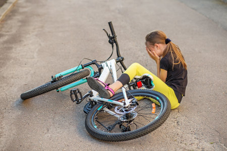 Young girl sits on ground, visibly upset, after falling from her bicycle. Her bike lies beside her on deserted street, showcasing moment of vulnerability and challenge.の写真素材