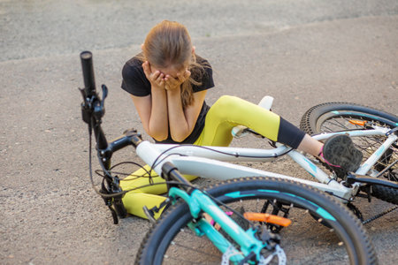 A young girl wearing a black shirt and yellow leggings sits on the ground, covering her face with her hands, clearly upset after falling off her bike. The bicycle lies beside her on the pavement.の写真素材