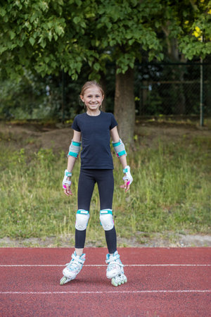 A preteen girl enjoys roller skating at an outdoor track. Wearing protective gear, she smiles...