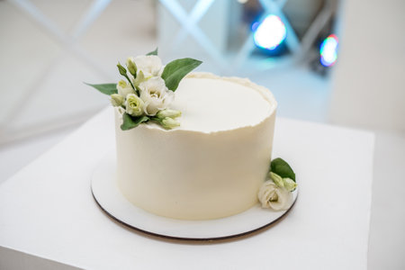 A minimalist white wedding cake with a smooth buttercream finish, adorned with delicate white flowers and greenery, displayed on a table.の写真素材