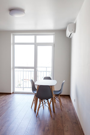 A bright and airy dining area features a sleek white table accompanied by four stylish chairs. Large windows let in natural light, creating a welcoming atmosphere in the apartment.の写真素材